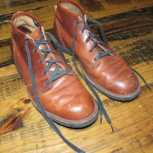May include: A pair of brown leather ankle boots with black laces. The boots have a worn appearance and are set on a wooden surface. The laces are untied, and the boots appear to be well-used.