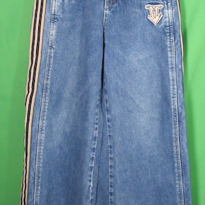 Könnte beinhalten: Blaue Jeans mit weitem Bein. Die Jeans haben einen dekorativen Streifen an der Seite mit schwarzen, beigen und weißen Streifen. Ein kleiner Aufnäher mit einem Logo befindet sich auf der Vordertasche.