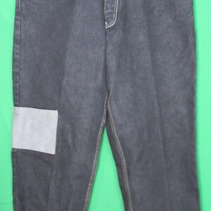 Puede incluir: Vaqueros de mezclilla de lavado oscuro con costuras blancas y parches. Presentan el texto "NATIONAL" en los bolsillos delanteros y un parche en la parte inferior de la pierna. Los vaqueros tienen un corte relajado y cuelgan de una percha blanca.