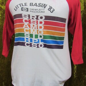 May include: White and red raglan-style t-shirt with the text "LITTLE BASIN '83" and "Hewlett Packard" printed on the front. The shirt features a rainbow-colored stripe design with various acronyms. The sleeves are red.