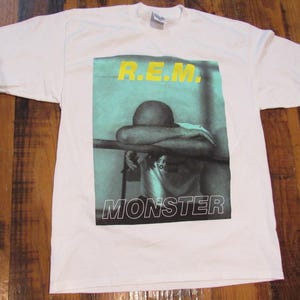 以下が含まれることがあります： 顔の前に腕を組んだ人物のグラフィックプリントが施された白いTシャツ。グラフィックは青と緑の色合いで、「R.E.M.」と「MONSTER」の文字が黄色で書かれています。