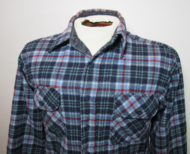 May include: A blue, green, and red plaid flannel shirt with two chest pockets and button fastenings. The shirt has a quilted collar and is displayed on a mannequin.