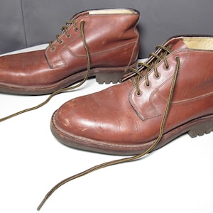 May include: A pair of brown leather ankle boots with matching laces. The boots have a classic design with a rounded toe and a sturdy sole. The laces are brown and yellow. The boots are placed on a white surface.