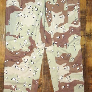 Könnte beinhalten: Wüsten-Tarnhose mit Knopfverschluss, Gürtelschlaufen und mehreren Taschen. Die Hose hat ein Muster aus unregelmäßigen Formen in Beige, Braun und Weiß. Die Hose hat Kordelzüge am unteren Ende der Beine.