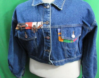 Peut inclure: Veste en jean bleue avec des boutons argentés et deux poches avant. La poche gauche a un patch coloré. La veste a des patchs décoratifs avec des ornements métalliques sur le côté gauche.