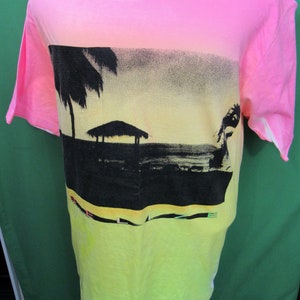May include: A neon pink and yellow t-shirt with a tropical beach scene. The top of the shirt and sleeves are pink, transitioning to yellow at the bottom. The graphic features palm trees, a hut, and a sunset.