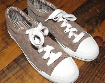 May include: A pair of brown suede sneakers with white laces and toe caps. The interior lining features a plaid pattern. The shoes are displayed on a wooden surface.
