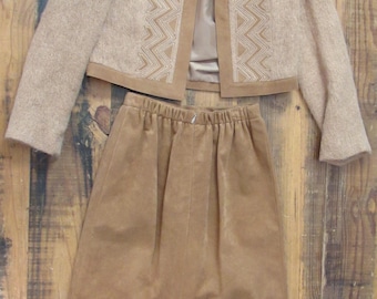 May include: A two-piece tan suit featuring a short jacket and a knee-length skirt. The jacket has a decorative geometric pattern and a contrasting trim. The skirt has an elastic waistband. The suit is displayed on a wooden surface.