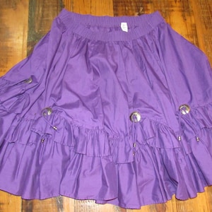 May include: A vibrant purple skirt with a ruffled hem and decorative silver-toned accents. The skirt features a gathered waistband and a flowing, flared design. The fabric has a slight sheen, adding to its visual appeal.