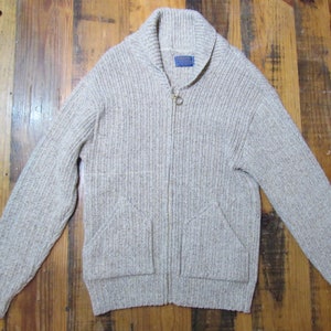 May include: A light brown, zip-up cardigan sweater with a shawl collar and two front pockets. The sweater has a ribbed texture and a small blue tag near the collar. The zipper is gold-colored.