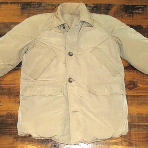 Puede incluir: Un abrigo de invierno acolchado de color beige con cierre de botones, dos bolsillos en el pecho y dos bolsillos inferiores. El abrigo tiene cuello y mangas largas. El abrigo está sobre una superficie de madera.