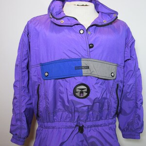 Puede incluir: Chaqueta anorak Ocean Pacific morada con capucha, cremalleras negras y cintura con cordón. La chaqueta tiene un bolsillo azul y gris con el logo de Ocean Pacific. La chaqueta se muestra en un maniquí blanco.