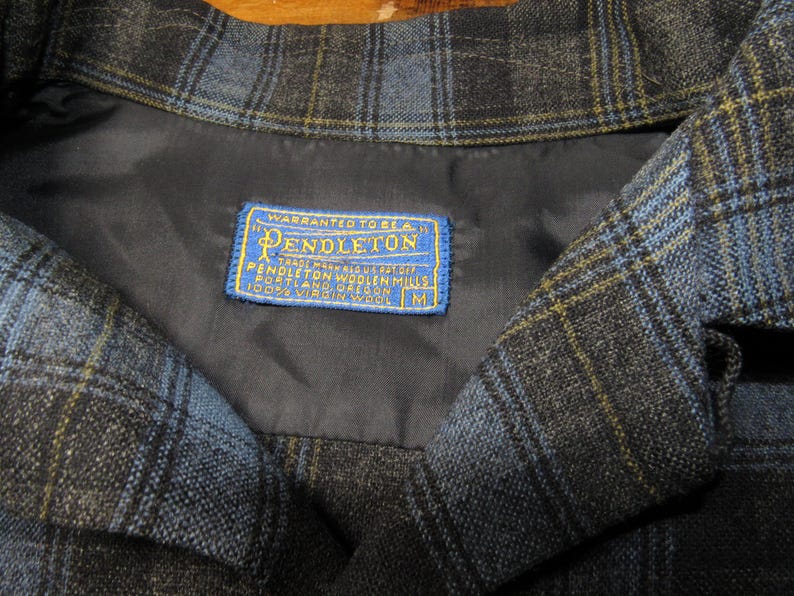 K&ouml;nnte beinhalten: Nahaufnahme eines Kragens einer blau, schwarz und gelb karierten Jacke. Die Jacke hat ein dunkelblaues Futter und ein blau-gelbes Etikett mit der Aufschrift "Pendleton" und "100% Virgin Wool". Die Jacke hat die Gr&ouml;&szlig;e M.