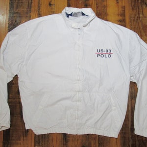 May include: White zip-up jacket with a stand-up collar. The jacket features the text "US-93 POLO" in red and navy blue on the chest. It has long sleeves and two front pockets. The jacket is made of a lightweight material.
