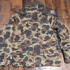 Puede incluir: Chaqueta de camuflaje con capucha, en tonos marrones, beige y verdes. Cuenta con múltiples bolsillos, cierre de botones y cordón en la cintura. Ideal para actividades al aire libre.