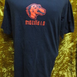 Pode incluir: Camiseta preta com um gráfico de dinossauro vermelho e o texto "mozilla 1.0" impresso na frente.