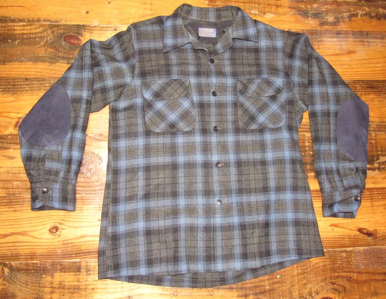 K&ouml;nnte beinhalten: Ein lang&auml;rmeliges kariertes Hemd in Blau-, Gr&uuml;n- und Graut&ouml;nen mit zwei Brusttaschen und dunkelblauen Ellbogenpatches. Das Hemd hat eine Knopfleiste und Manschetten.