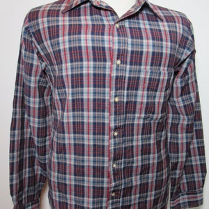 May include: A long-sleeved plaid shirt in a classic style. The shirt features a pattern of navy blue, red, and light blue checks. It has a button-down front, a chest pocket, and a collared neckline. The shirt is displayed on a mannequin.
