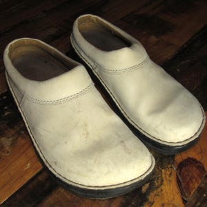 May include: A pair of off-white leather clogs with black soles. The clogs have a rounded toe and a slip-on design. The stitching is visible around the edges of the shoe. The shoes appear worn.