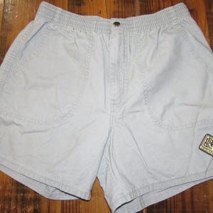 May include: Pale grey shorts with an elasticated waistband and button fastening. The shorts feature two front pockets and a small square patch on the right leg. They are made from a lightweight material.