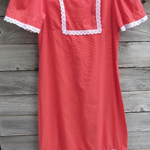 May include: A vintage red dress with white polka dots and white lace trim. The dress has short sleeves, a square neckline, and a ruffled hem. It hangs on a wooden hanger against a wooden background.