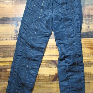 May include: Black nylon pants with multiple zipper details. The pants have a drawstring waistband and a relaxed fit.