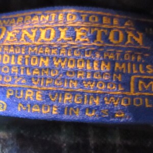 May include: A close-up of a blue and gold Pendleton label. The label reads "WARRANTED TO BE A PENDLETON" and includes text about the brand, location, and material. The label is stitched with a white zig-zag pattern and has a size "M".
