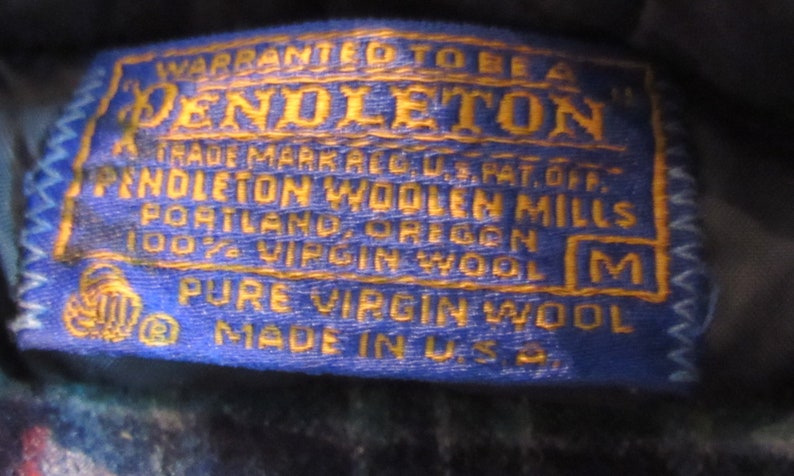 May include: A close-up of a blue and gold Pendleton label. The label reads "WARRANTED TO BE A PENDLETON" and includes text about the brand, location, and material. The label is stitched with a white zig-zag pattern and has a size "M".