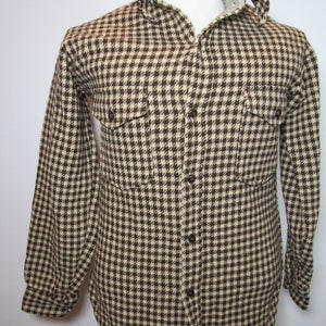 Può includere: Una camicia a maniche lunghe con un motivo a pied-de-poule marrone e crema. La camicia ha una chiusura con bottoni sul davanti, due tasche sul petto e un colletto. Il colletto è foderato con un tessuto colorato chiaro. La camicia è esposta su un manichino.