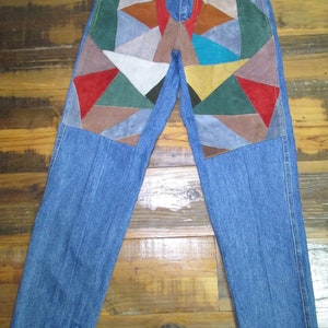May include: Blue denim jeans with colourful geometric suede patches on the front. The patches feature triangles in shades of red, brown, green, and blue. The jeans have a classic five-pocket design.