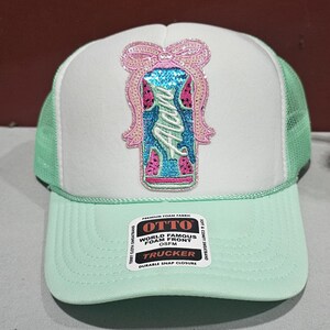 K&ouml;nnte beinhalten: Eine mintgr&uuml;ne und wei&szlig;e Trucker-M&uuml;tze mit einem Pailletten-Design. Das Design zeigt eine Schleife und das Wort "Alana" in einer dekorativen Schriftart. Die M&uuml;tze hat eine Mesh-R&uuml;ckseite und einen Schnappverschluss.