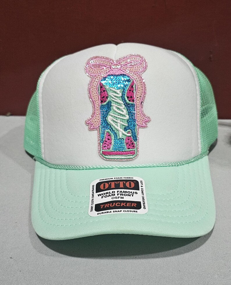 K&ouml;nnte beinhalten: Eine mintgr&uuml;ne und wei&szlig;e Trucker-M&uuml;tze mit einem Pailletten-Design. Das Design zeigt eine Schleife und das Wort "Alana" in einer dekorativen Schriftart. Die M&uuml;tze hat eine Mesh-R&uuml;ckseite und einen Schnappverschluss.