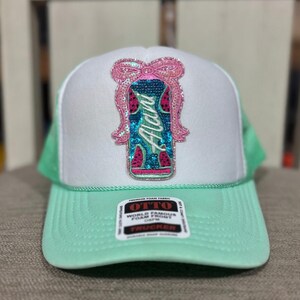 K&ouml;nnte beinhalten: Eine wei&szlig;e und mintgr&uuml;ne Trucker-M&uuml;tze mit Pailletten-Design. Das Design zeigt das Wort "Alani" in T&uuml;rkis, eine rosa Schleife und Wassermelonenst&uuml;cke. Die M&uuml;tze hat einen mintgr&uuml;nen Schirm und ein Mesh-R&uuml;ckenteil.
