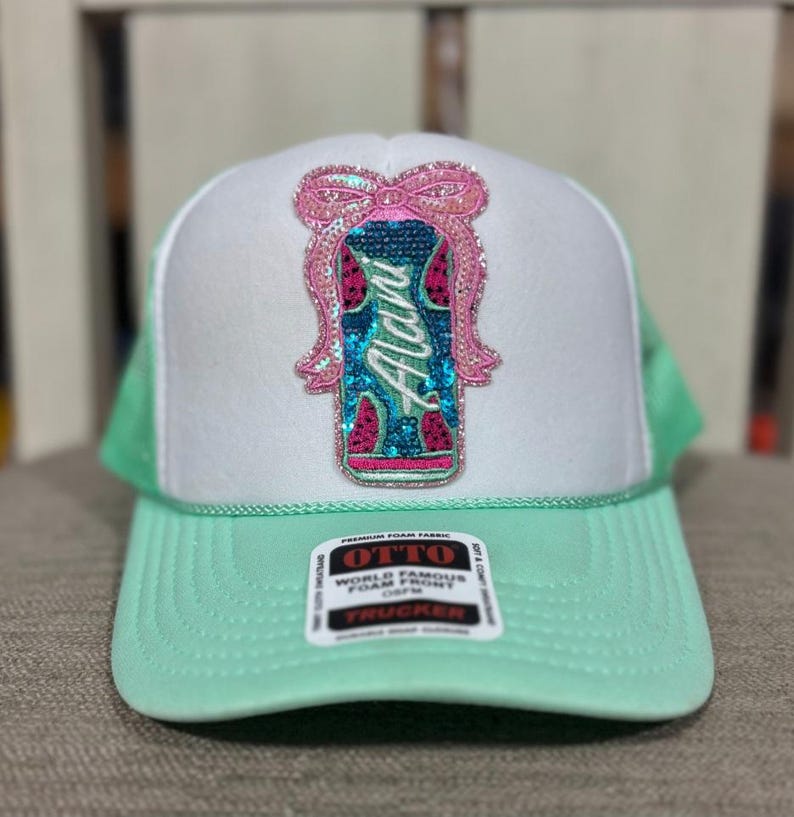 K&ouml;nnte beinhalten: Eine wei&szlig;e und mintgr&uuml;ne Trucker-M&uuml;tze mit Pailletten-Design. Das Design zeigt das Wort "Alani" in T&uuml;rkis, eine rosa Schleife und Wassermelonenst&uuml;cke. Die M&uuml;tze hat einen mintgr&uuml;nen Schirm und ein Mesh-R&uuml;ckenteil.