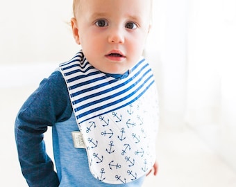 May include: A baby wearing a blue and white striped bib with a white background and blue anchor print. The bib has a blue trim around the edges.