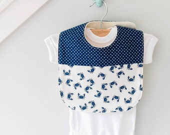 May include: A baby bib and onesie set. The bib features a navy blue polka dot pattern on top and a white background with blue crab illustrations on the bottom. The onesie is white and the set is hanging on a wooden hanger.
