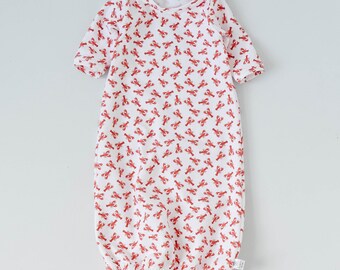 May include: White baby gown with a repeating pattern of red lobsters. The gown has long sleeves and a gathered elastic hem. The neckline is a simple round shape. The fabric appears to be soft and lightweight.