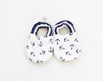 May include: A pair of white baby shoes with a navy blue anchor pattern. The shoes have navy blue and white striped trim and a soft, white interior. The shoes are designed for infants and toddlers.