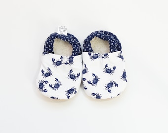 May include: A pair of white baby booties with a blue crab pattern. The booties are lined with a soft, white material.