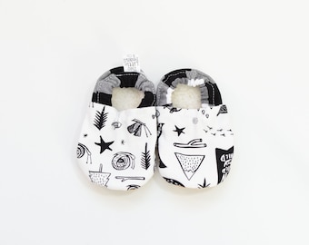 May include: A pair of baby shoes with a black and white pattern. The shoes have a white base with black illustrations of stars, trees, and other shapes. The shoe opening is gray and black checkered. The shoes are on a white background.