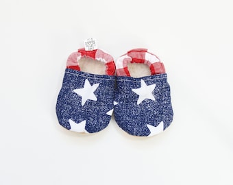 May include: A pair of baby moccasins with a patriotic theme. The shoes feature a blue denim-like fabric with white stars, red and white gingham trim, and a soft, cream-colored interior. The shoes are designed for infants.