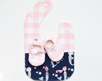 May include: A baby bib and matching shoes set. The bib features a pink and white gingham pattern on top, transitioning to a navy blue fabric with mermaid illustrations. The shoes are pink and white gingham with a soft, white interior.