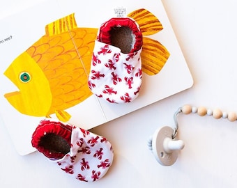 May include: A children's book with a picture of a goldfish and the text "Goldfish, Goldfish, What do you see?"  The book is open to a page with a picture of a teacher looking at the reader.  Two red and white baby shoes with a lobster design are on top of the book.  A wooden bead necklace with a white silicone pacifier is on the white surface.