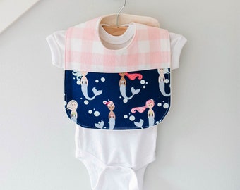 May include: A white baby onesie with a pink and white gingham bib featuring a mermaid print. The bib has a navy blue background with white bubbles and mermaids with pink, blue, and blonde hair.