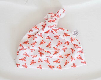 May include: White baby hat with a red lobster print and a knot on top. The hat has a small tag that says "The Little Things" on it.