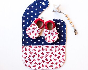 May include: A blue and white bib with stars and a red and white lobster print. Two red and white lobster print baby shoes are on top of the bib.