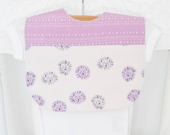 May include: A baby bib with a lavender top section featuring a white star pattern. The lower section is off-white and decorated with repeating lion face illustrations in lavender and navy blue. The bib is hanging on a wooden hanger.