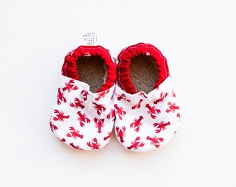 May include: A pair of red and white baby booties with a lobster print. The booties are made of soft fabric and have a red trim around the top.