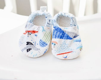 May include: A pair of baby shoes with a blue, white, and red nautical themed print. The shoes are made of soft fabric and have a white fleece lining. The shoes have a tag that says "Little Feet Only" and "Two Pairs".