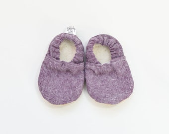 May include: A pair of purple baby moccasins with a textured fabric exterior and a soft, white interior. The shoes are designed with a rounded toe and a simple, comfortable design. A small tag is attached to one shoe.